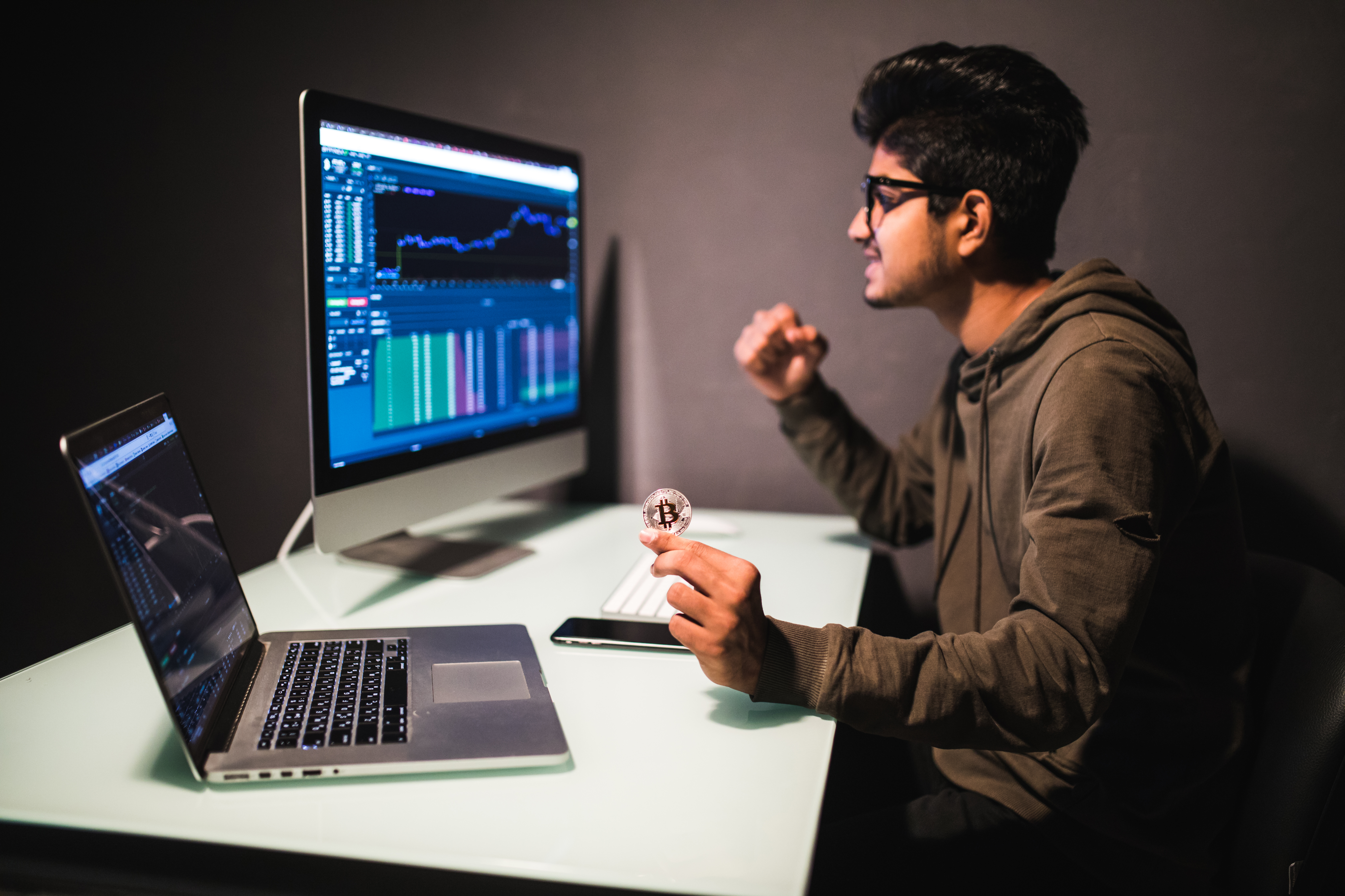 Focused trader analyzing market charts on dual screens while holding a Bitcoin coin, symbolizing disciplined and data-driven trading practice.